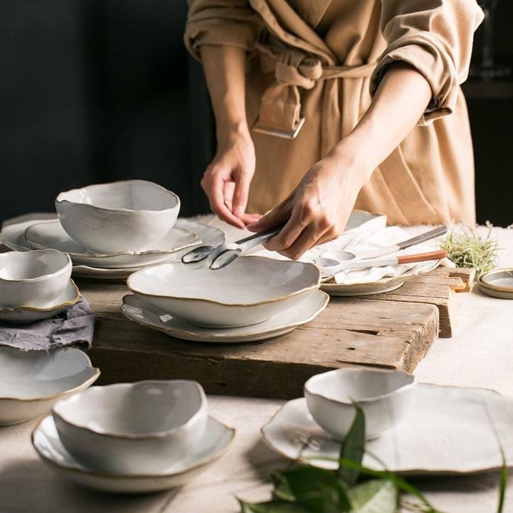 Woman arranging Nanna Bowls And Dishes from Thetis Tableware Set on wooden table; elegant ceramic kitchenware collection.