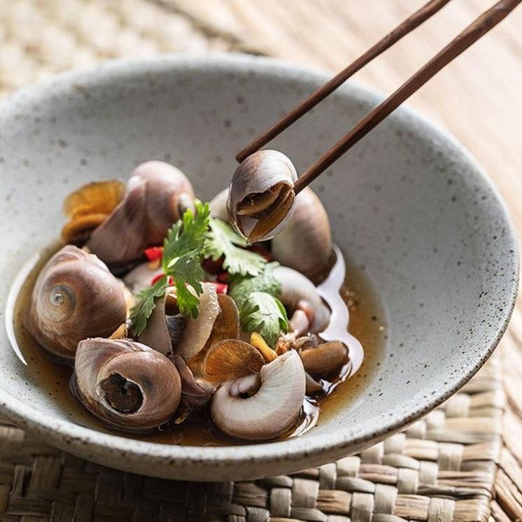 Chopsticks holding food over a Hemera Ceramic Bowl with snails and herbs
