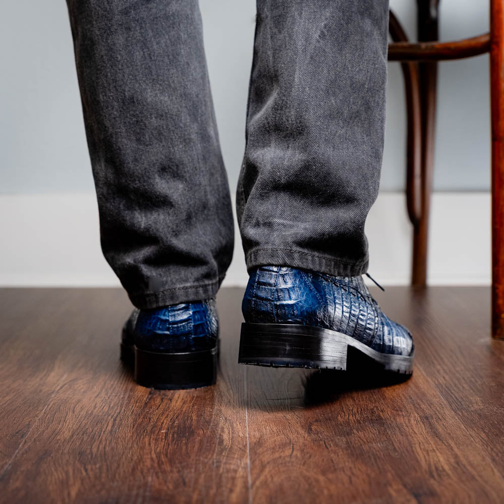 Back view of Ragusa Stained Navy shoes in genuine caiman skin with hybrid sole, worn with dark pants on wooden floor.