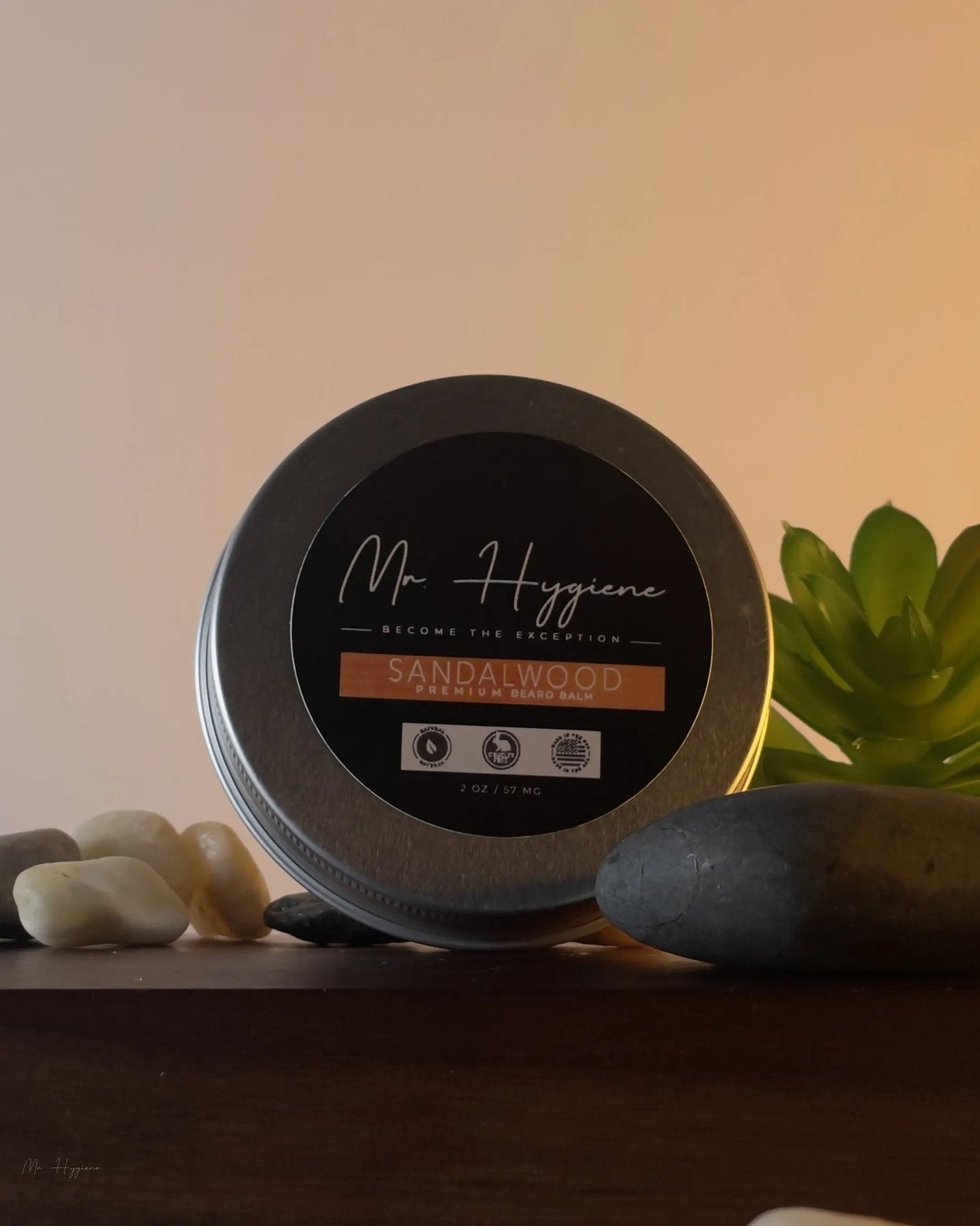 Tin of 'Sandalwood Beard Balm for Men – Shape, Soften & Scent' on a wooden shelf with pebbles and plant.