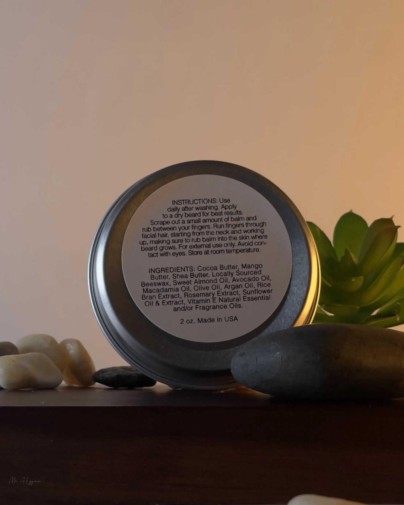 Back view of Sandalwood Beard Balm for Men – Shape, Soften & Scent, showing ingredients and instructions label, surrounded by natural stones.