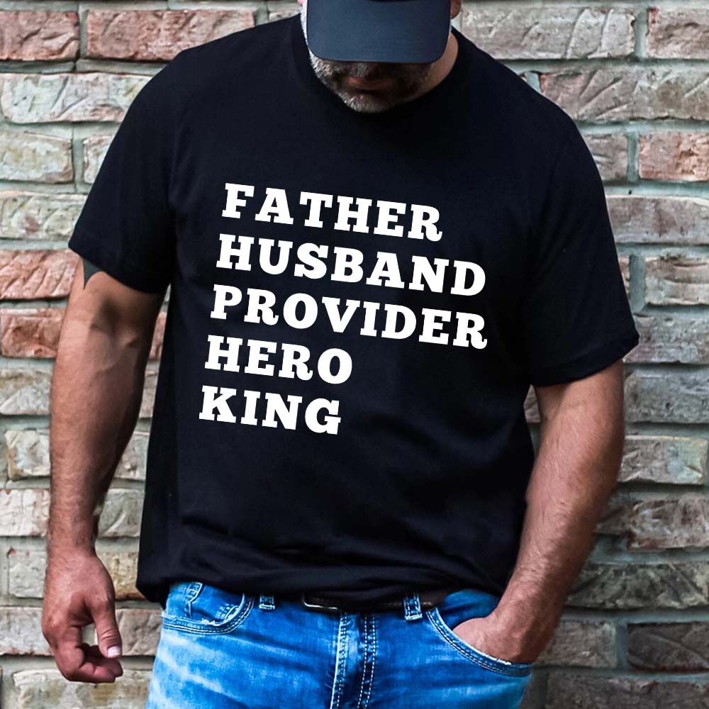 Man wearing Father Husband Provider Hero King T-shirt against brick wall.