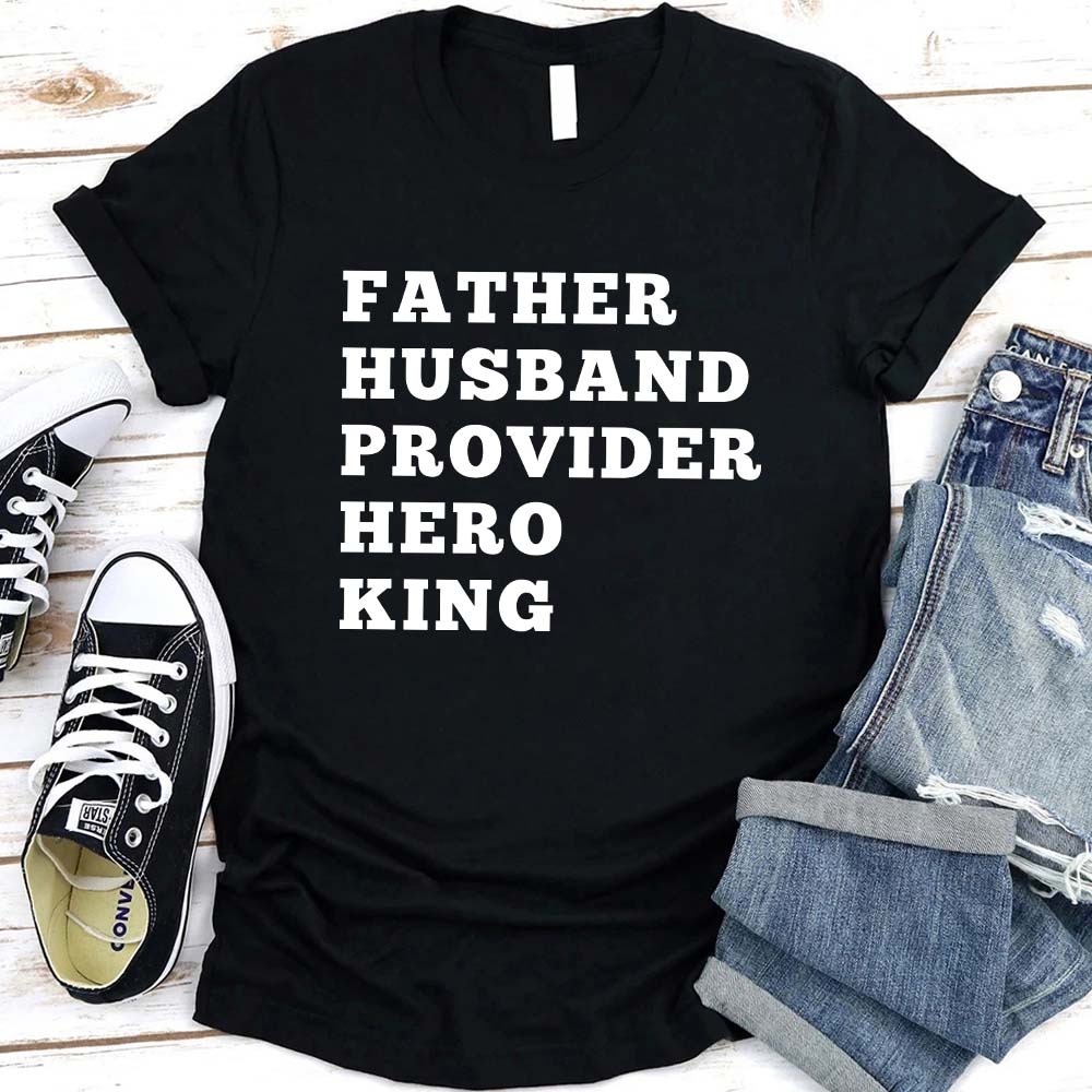 Father Husband Provider Hero King T-shirt with bold white lettering on black fabric, folded jeans, and sneakers on rustic background.