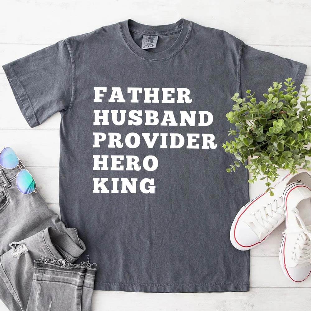 Father Husband Provider Hero King T-shirt with bold white text, surrounded by casual accessories like jeans, sunglasses, and sneakers.